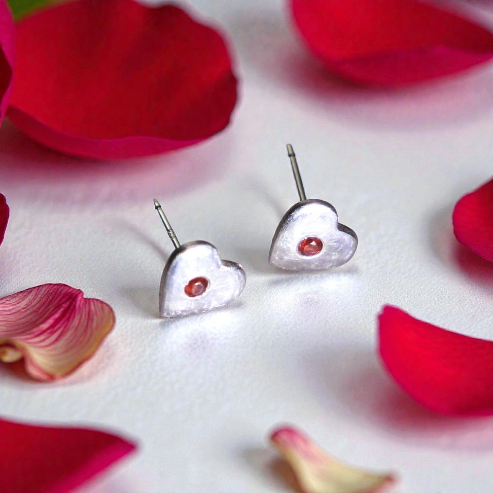 Two silver heart shaped stud earrings resting on their sides. They are sitting on a reflective white surface, surrounded by red rose petals.
