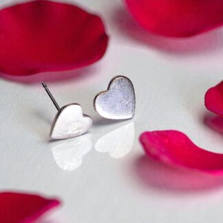 A pair of silver heart shaped stud earrings sitting on a white reflective surface. The shine from the silver is reflected in the surface. Surrounding the earrings are five red rose petals.