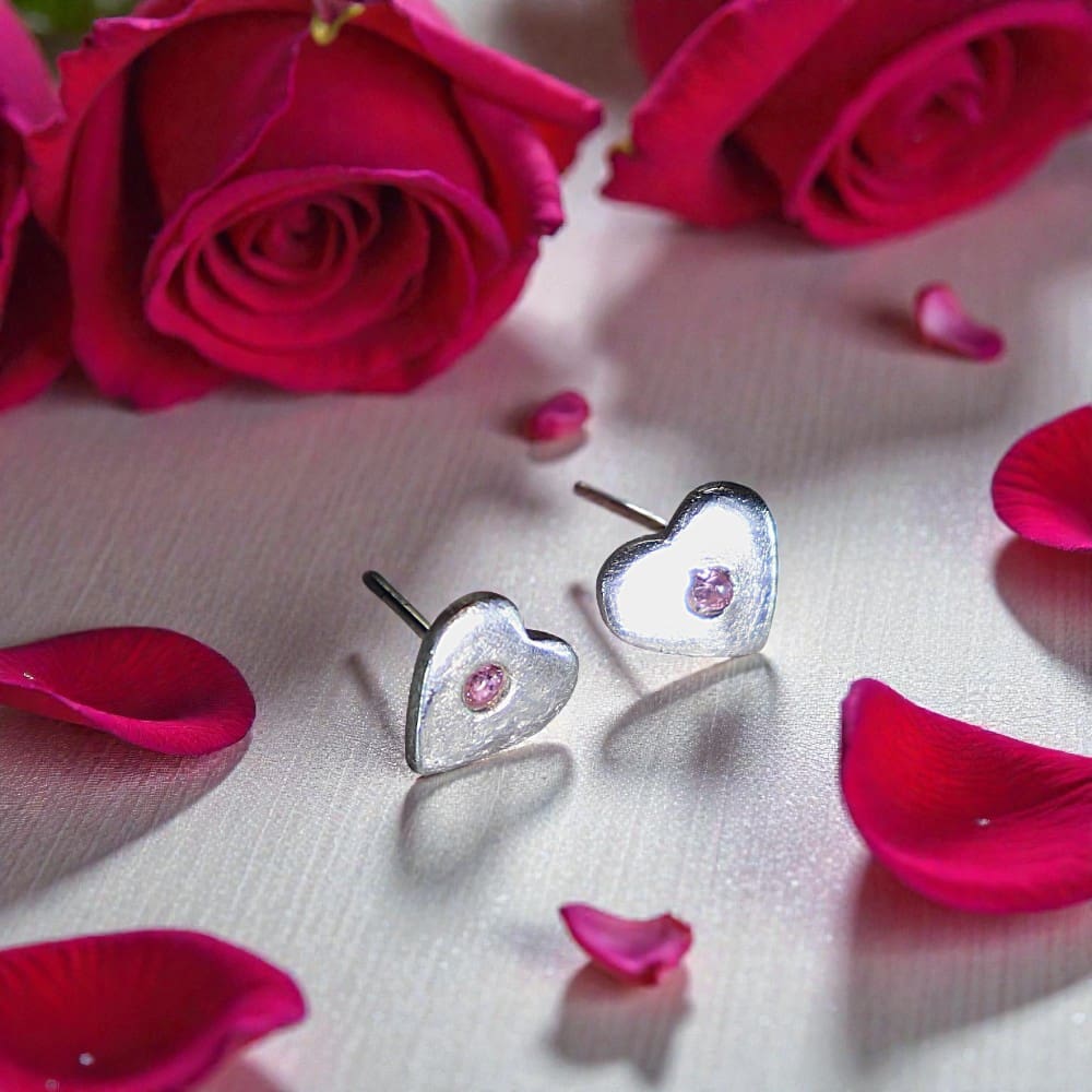 A pair of silver, heart shaped studs on a white reflective surface. Each heart has a pink stone in the middle. Behind the earrings are two red roses and to the front and either side of them are red rose petals