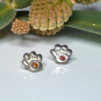 A pair of silver paw print shaped stud earrings with red stones in the middle are sitting on a shiny white surface. In the background behind the earrings are a some green leaves and a brown seed