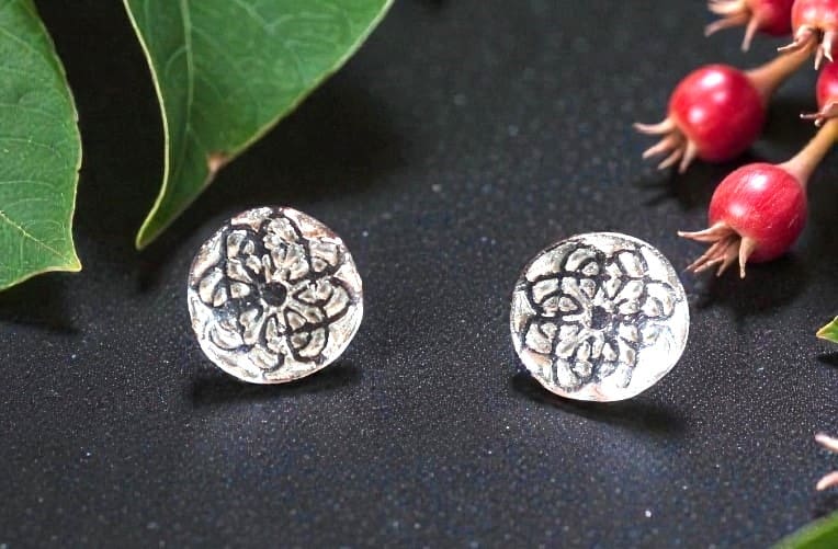 Two round silver studs inlaid with a comlex Celtic Knot design. They are sitting on a matt black counter top. To the top left are two large green leaves. To the top right are two red rosehip buds