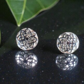 Two round silver studs with a design of several overlaid Celtic Knots. They are sitting on a reflective black counter top, their reflection can be seen in it. Behind and to the left are two Montesera leaves