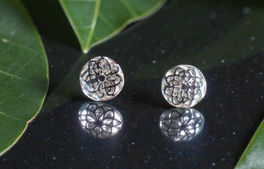 Two round silver studs with a design of several overlaid Celtic Knots. They are sitting on a reflective black counter top, their reflection can be seen in it. Behind and to the left are two Montesera leaves