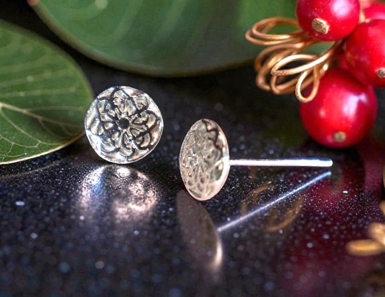 Two round silver studs on a shiny back countertop. One stud is facing forward whilst the other one is at angle so that the post can be seen behind it. Behind the first stud are two large green leaves. At the top right of the picture almost touching the post of the second stud are three red seeds