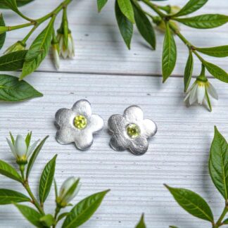A pair of silver daisy shaped earrings, with a yellow gem in the middle of each of them. They are sitting on a white painted wooden tables framed by four white flowers on long stems with long green leaves.