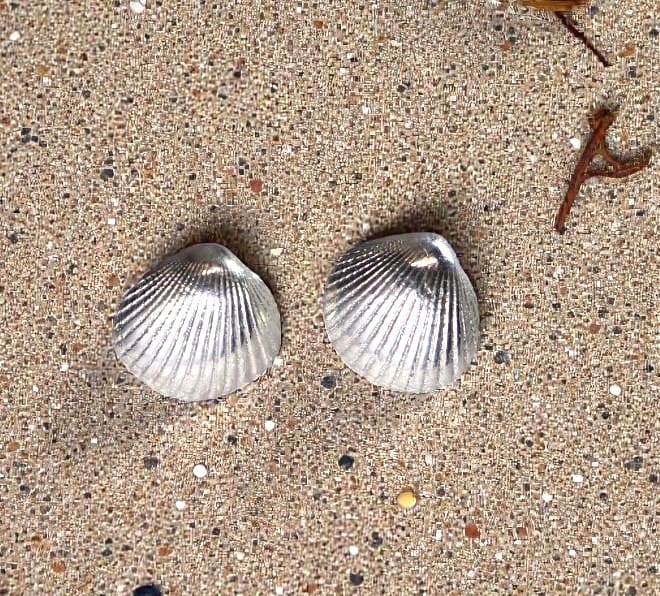 Silver seashell studs sitting on some sand with small bits of seaweed to the top right of the picture