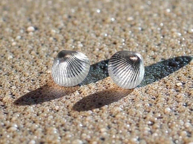 Silver cockleshell stud earrings resting on sand with their shadows going out in two directions