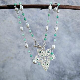 Etched sterling silver ivy pendant set with white sapphires, tsavorite garnets and lab emeralds, strung on a silk necklace with emerald, labradorite and baroque pearls. Hanging on a grey background. Isobel Kelly Jewellery, Hants UK