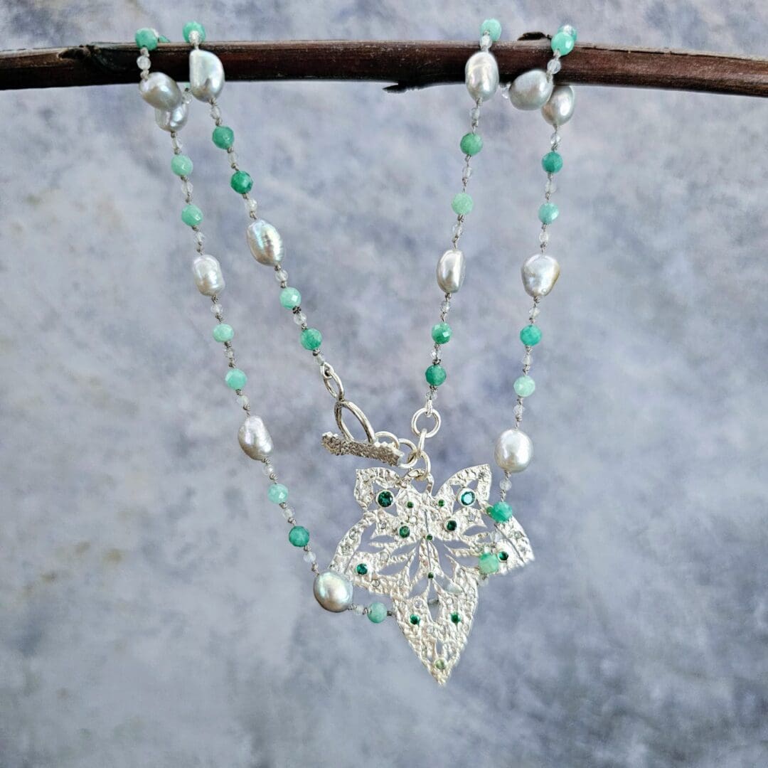 Etched sterling silver ivy pendant set with white sapphires, tsavorite garnets and lab emeralds, strung on a silk necklace with emerald, labradorite and baroque pearls. Hanging on a grey background. Isobel Kelly Jewellery, Hants UK