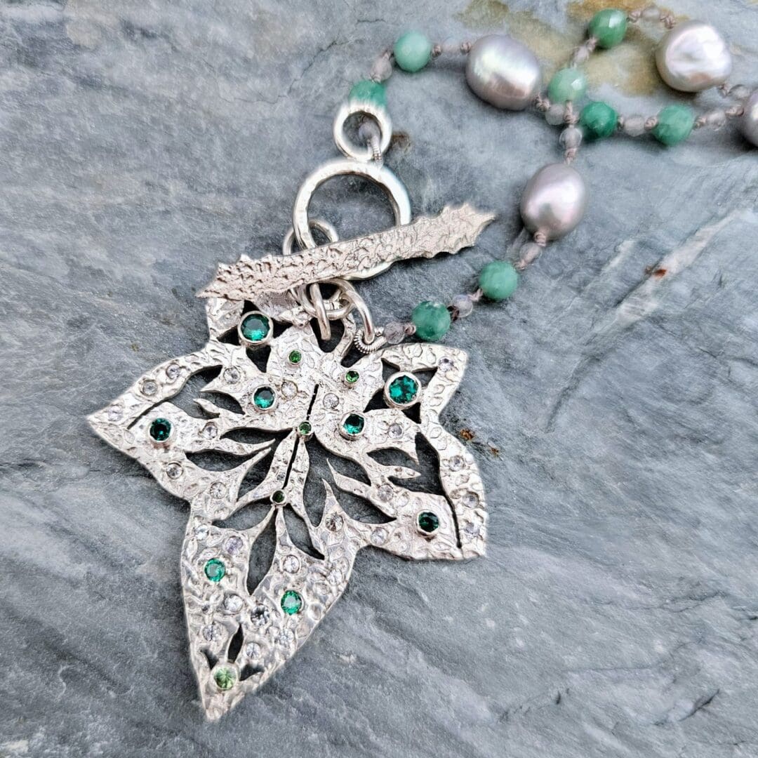 Etched sterling silver ivy pendant set with white sapphires, tsavorite garnets and lab emeralds, strung on a silk necklace with emerald, labradorite and baroque pearls. Flat. Isobel Kelly Jewellery, Hants UK