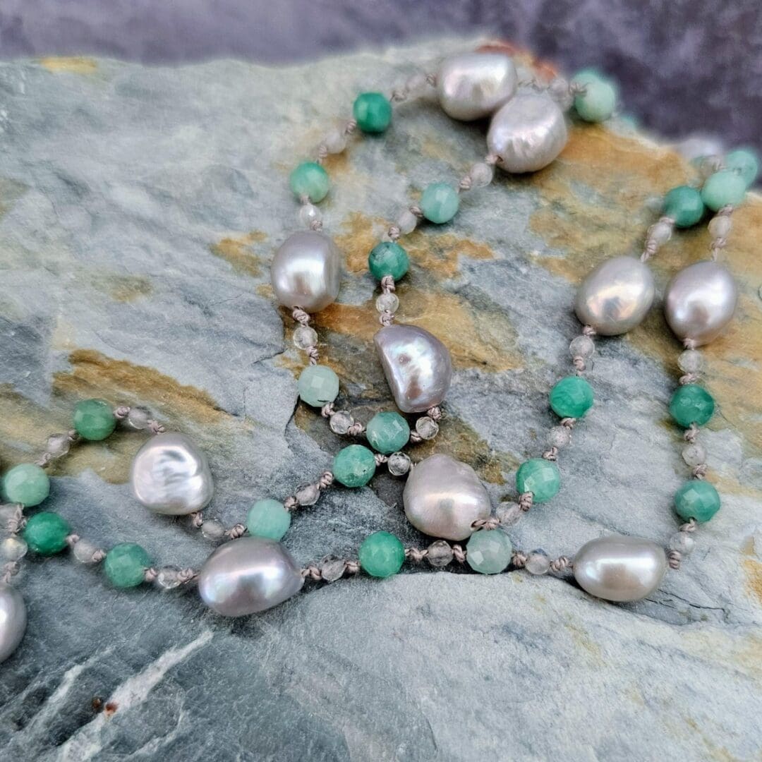 Etched sterling silver ivy pendant set with white sapphires, tsavorite garnets and lab emeralds, strung on a silk necklace with emerald, labradorite and baroque pearls. Bead close up on rock. Isobel Kelly Jewellery, Hants UK