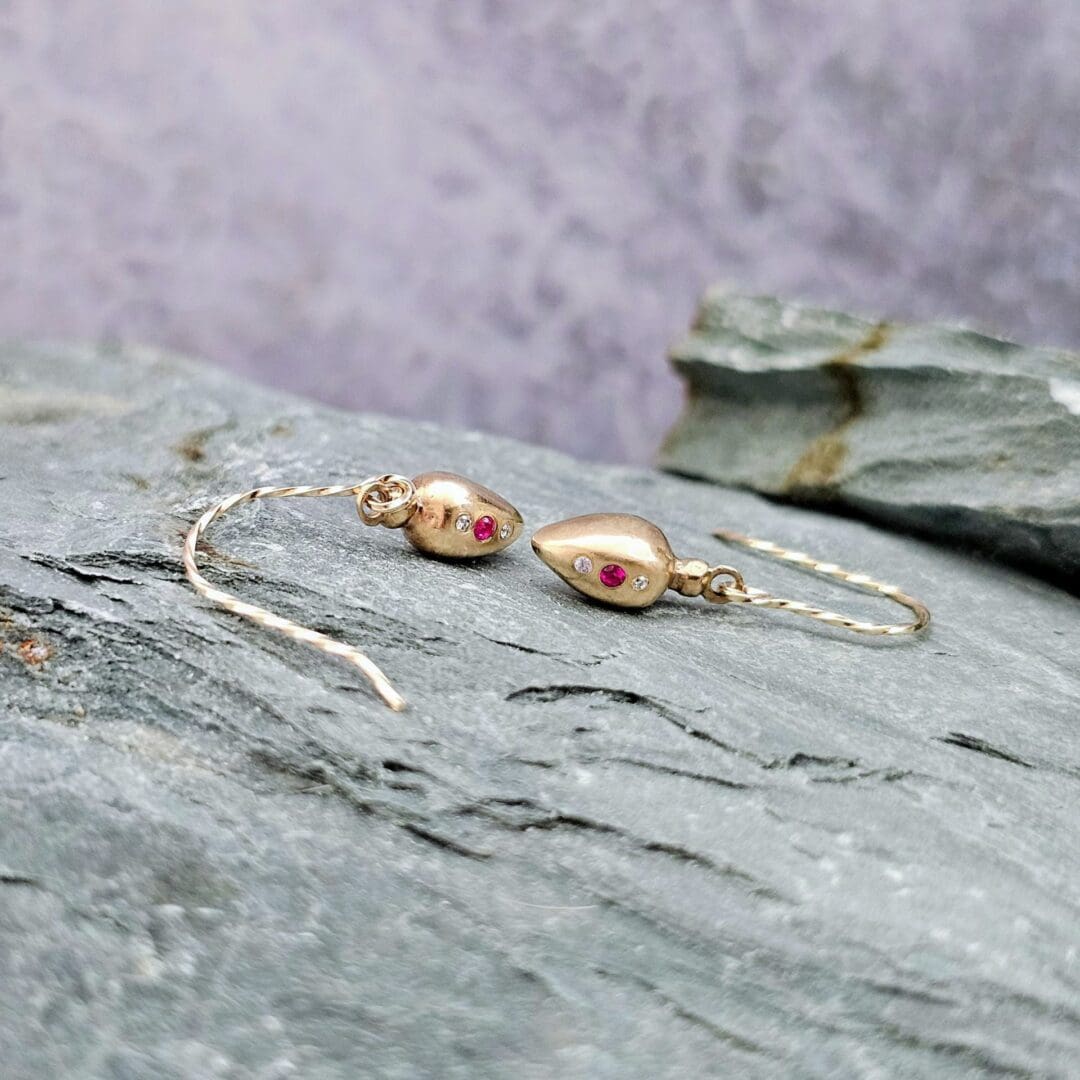 9ct gold drop earrings inspired by Roman amphora vessels, set with lab-grown diamonds and a central pink ruby, featuring twisted gold hooks. Side profile, on grey rock. Isobel Kelly Jewellery, Hants UK