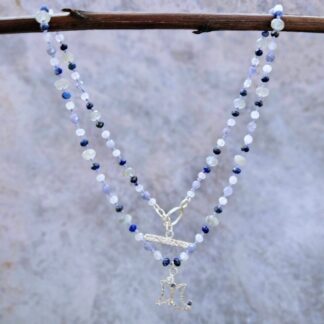 Silver triple moon pendant set with tiny gemstones, on a knotted silk strand of faceted blue, white and grey beads, finished with a textured toggle clasp. Hanging next to grey background. Isobel Kelly Jewellery, Hants UK