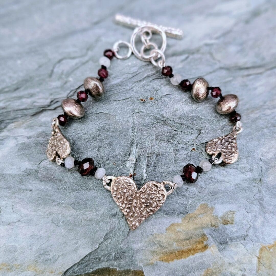 Handcrafted silver heart bracelet with antiqued etched charms, red garnet and moonstone beads on knotted silk, finished with a toggle clasp, Isobel Kelly Jewellery, Hants UK
