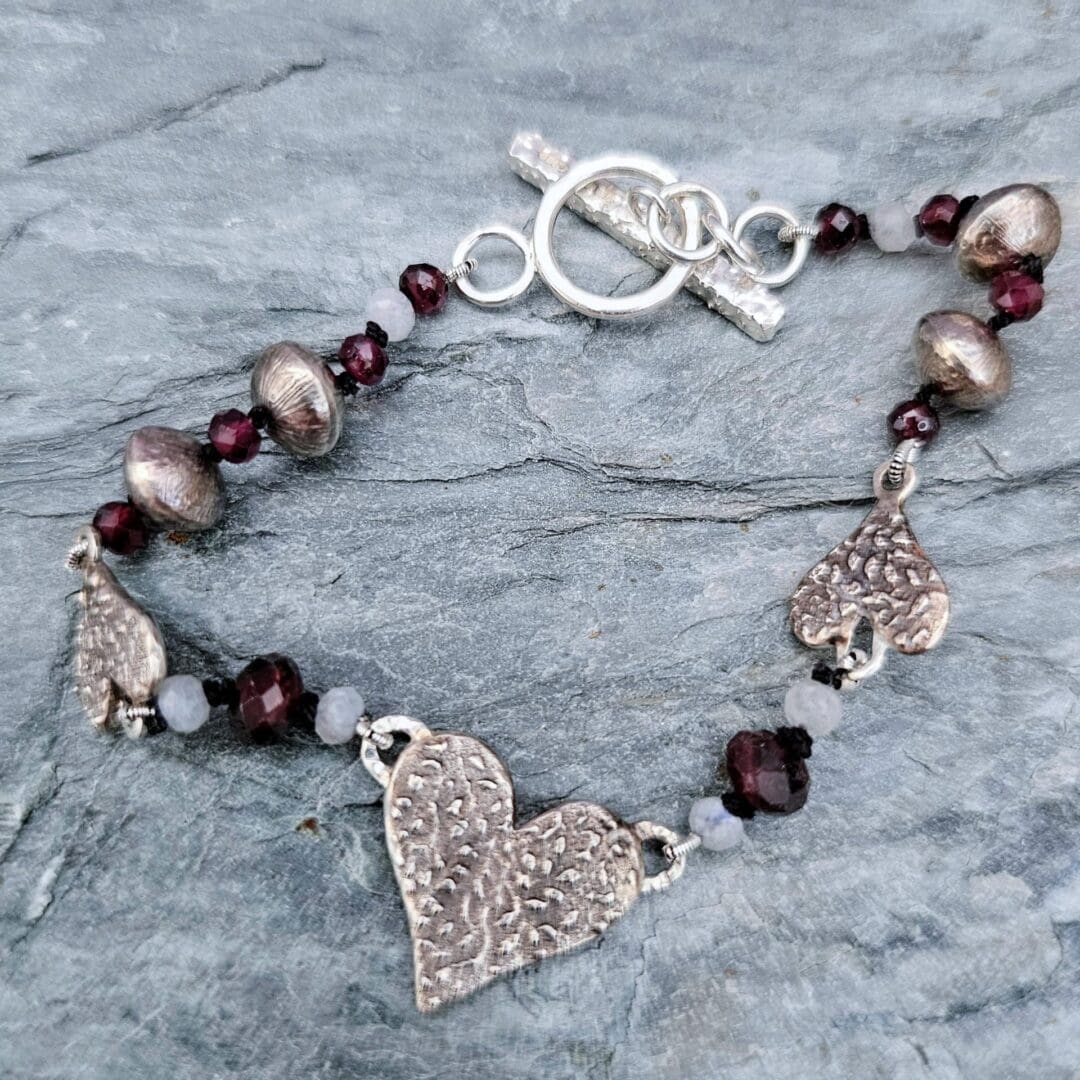 Handcrafted silver heart bracelet with antiqued etched charms, red garnet and moonstone beads on knotted silk, finished with a toggle clasp, grey background, Isobel Kelly Jewellery, Hants UK