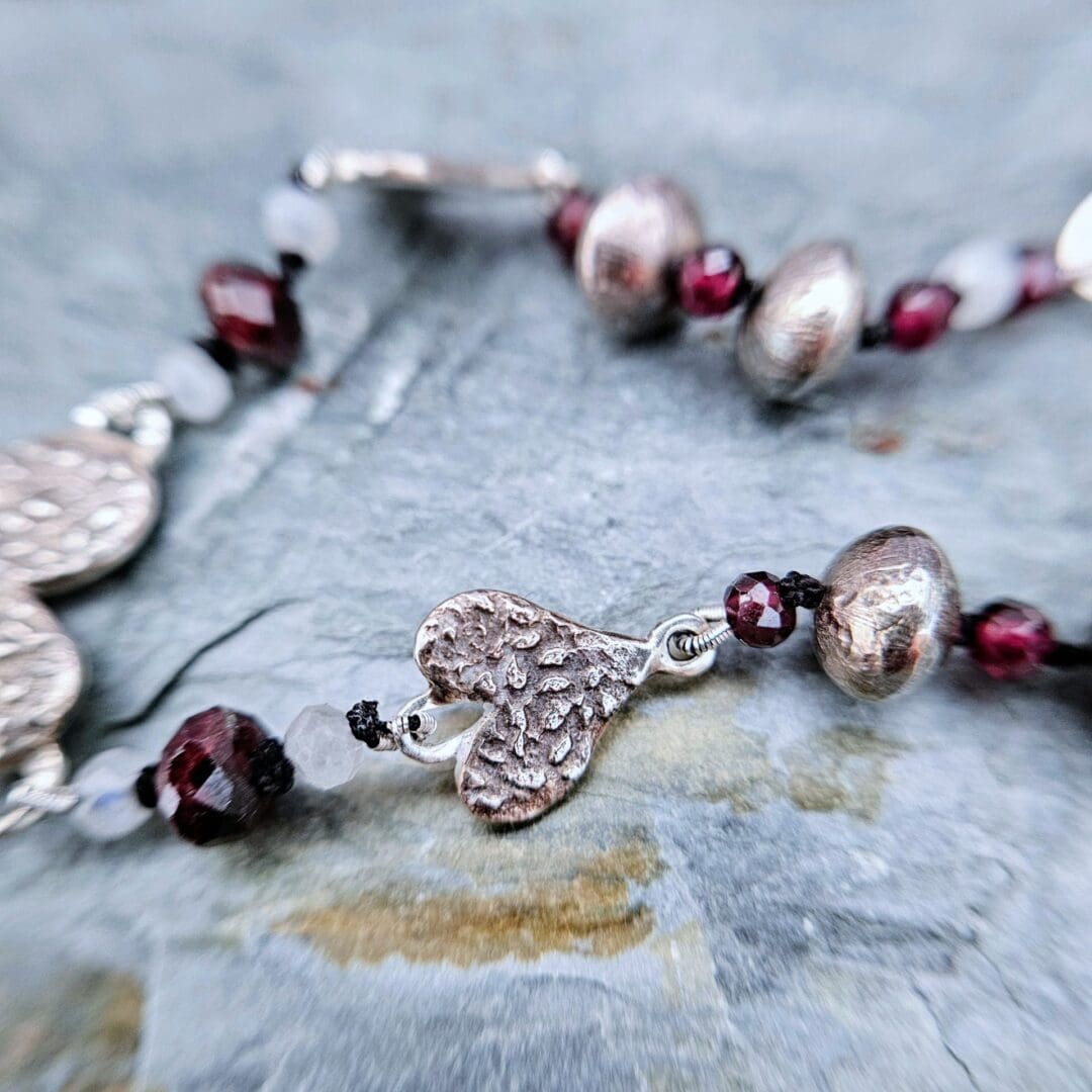 Handcrafted silver heart bracelet with antiqued etched charms, red garnet and moonstone beads on knotted silk, finished with a toggle clasp, heart and bead detail, Isobel Kelly Jewellery, Hants UK
