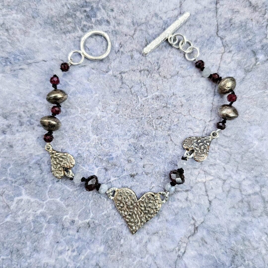 Handcrafted silver heart bracelet with antiqued etched charms, red garnet and moonstone beads on knotted silk, finished with a toggle clasp, flat grey stone shot, Isobel Kelly Jewellery, Hants UK
