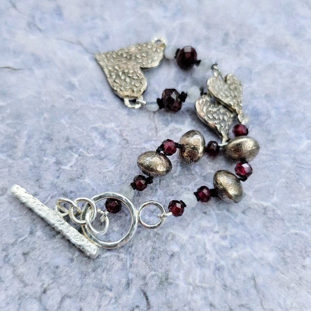 Handcrafted silver heart bracelet with antiqued etched charms, red garnet and moonstone beads on knotted silk, finished with a toggle clasp, toggle and bead detail, Isobel Kelly Jewellery, Hants UK
