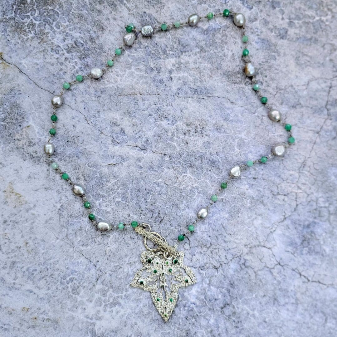 Etched sterling silver ivy pendant set with white sapphires, tsavorite garnets and lab emeralds, strung on a silk necklace with emerald, labradorite and baroque pearls. Flat grey background. Isobel Kelly Jewellery, Hants UK