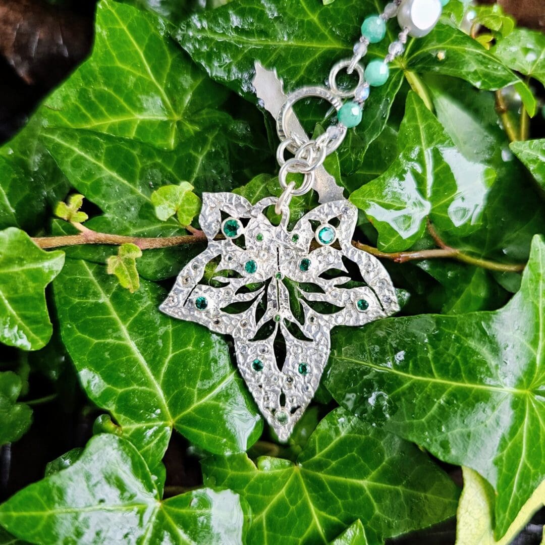 Etched sterling silver ivy pendant set with white sapphires, tsavorite garnets and lab emeralds, strung on a silk necklace with emerald, labradorite and baroque pearls. Ivy pendant amongst ivy. Isobel Kelly Jewellery, Hants UK
