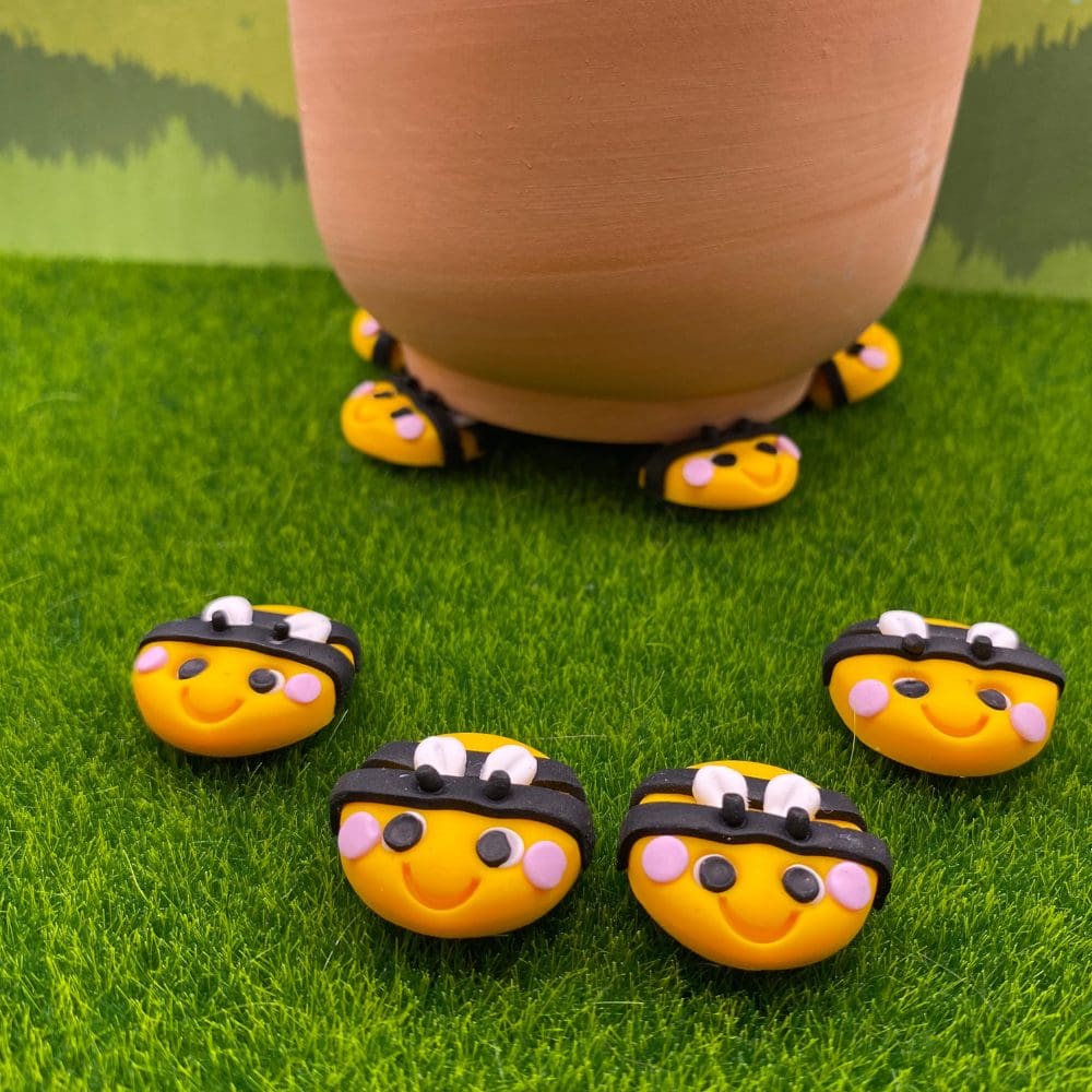 A terracotta pot rests on yellow and black pot feet, with four more bee decorations scattered on the artificial grass in front of it, which shows there smiling faces and small white wings.