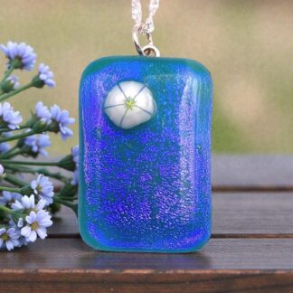 A vibrant blue glass pendant with a white flower design, resting beside delicate blue flowers on a wooden surface.