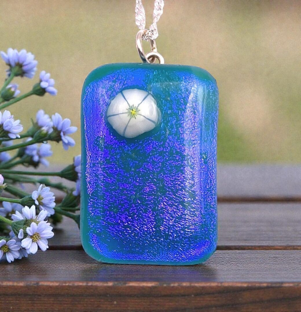 A vibrant blue glass pendant with a white flower design, resting beside delicate blue flowers on a wooden surface.