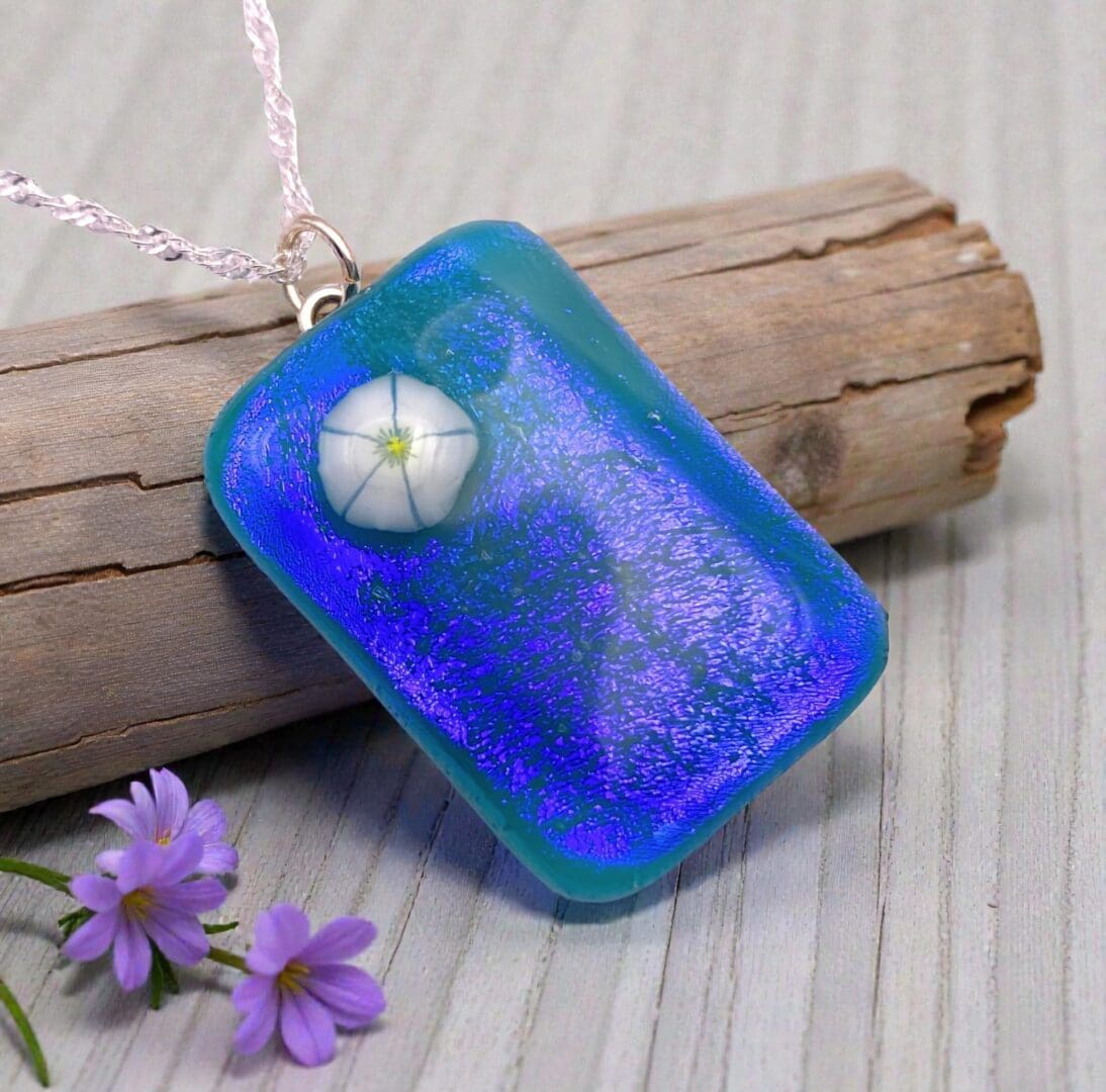 A vibrant blue pendant with a floral design, resting on a weathered wood surface, alongside delicate purple flowers.