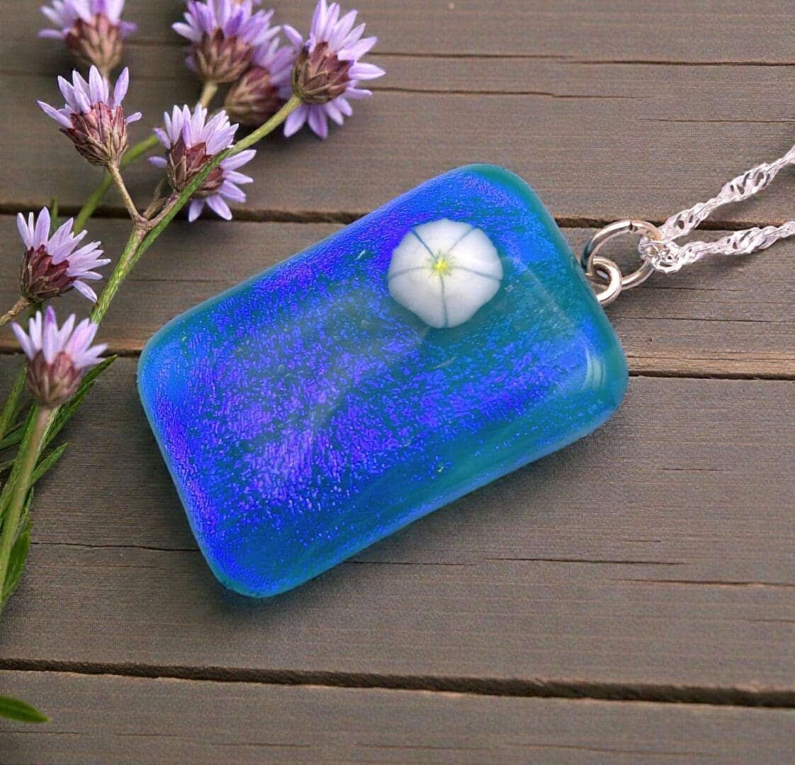 A blue and purple pendant featuring a small white flower, resting on a wooden surface alongside delicate purple flowers.