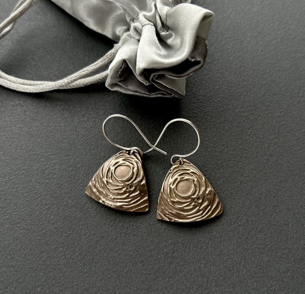 A pair of handcrafted bronze triangular drop earrings that have a swirl textured raised pattern. They have sterling silver ear wires and are displayed on a dark grey background.