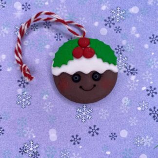 A Christmas pudding ornament with a smiling face, red holly berries, and a white icing trim, hangs from a red and white twine loop against a purple background with white snowflakes.