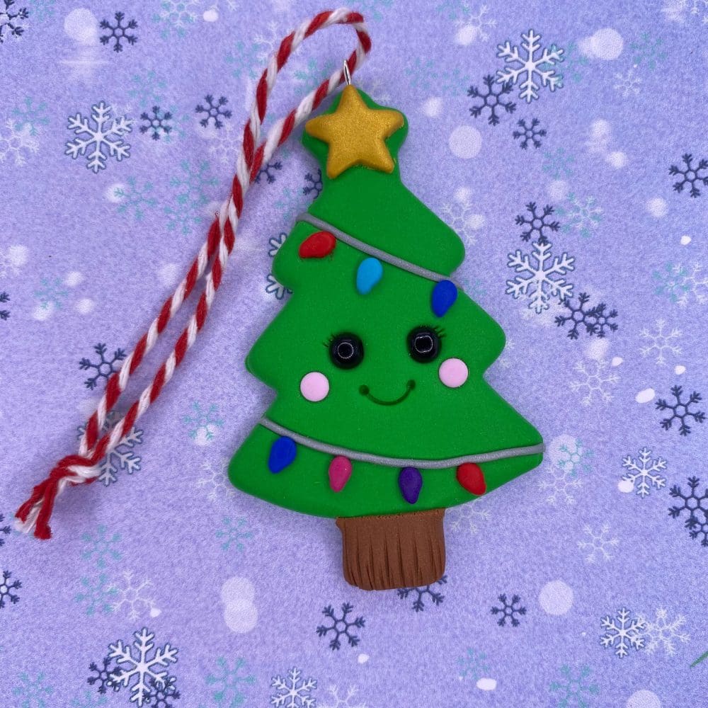 The picture is of a handmade Christmas tree polymer clay decoration with a star on top, a smiling face and strings of multi-coloured lights including black bead eyes, a smiling face and rosey cheeks. with a red and white string to hand on the tree. On a lilac background with snowflakes in differing size and colours.