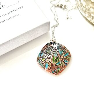 A handcrafted copper pendant in a soft diamond shape and a textured pattern that has been hand-painted in turquoise and green patina paints. The pendant has a sterling silver chain and is displayed on a white surface.