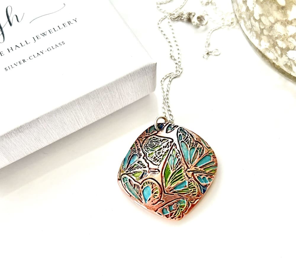 A handcrafted copper pendant in a soft diamond shape and a textured pattern that has been hand-painted in turquoise and green patina paints. The pendant has a sterling silver chain and is displayed on a white surface.