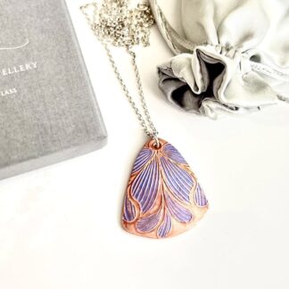 A handcrafted copper pendant which is textured with a flower pattern hand painted in lilac metallic paints. It has a sterling silver chain and is displayed on a white background.
