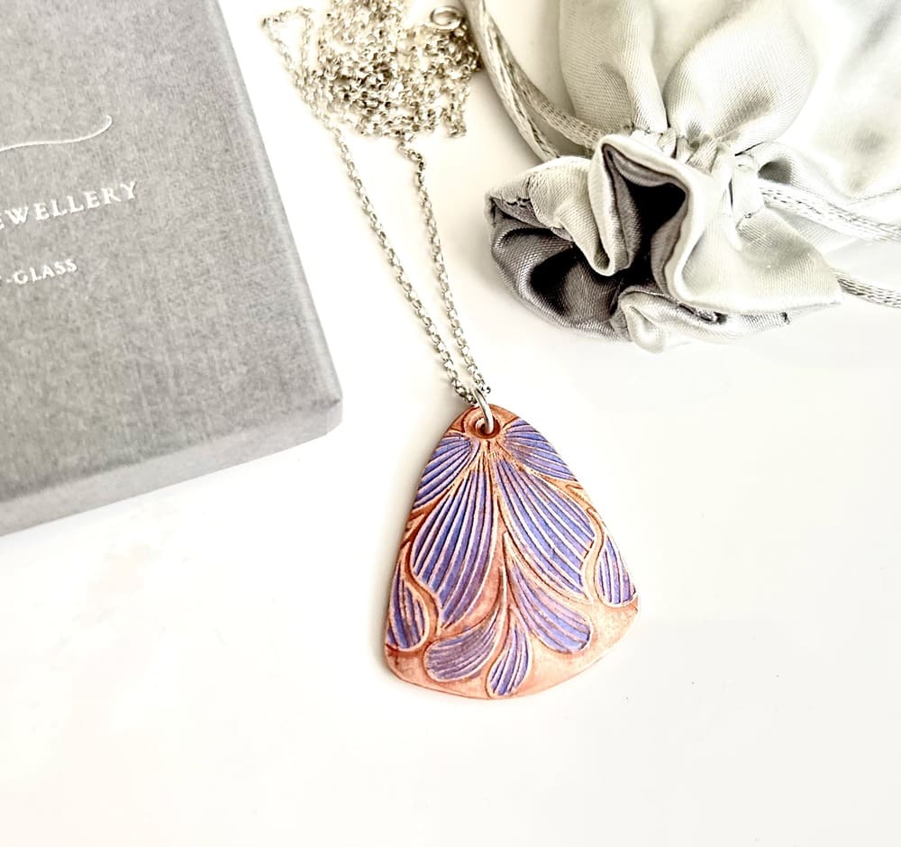 A handcrafted copper pendant which is textured with a flower pattern hand painted in lilac metallic paints. It has a sterling silver chain and is displayed on a white background.