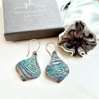 A pair of handcrafted copper drop earrings in a round shape with a swirl textured pattern. They are hand-painted in soft aqua and pale turquoise and have copper ear sires, displayed on a white background with a grey gift box and satin jewellery pouch above them.