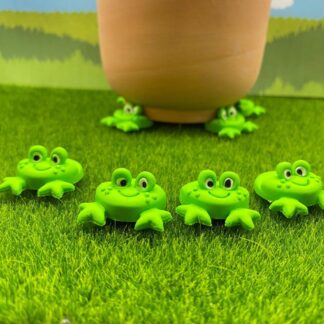 A terracotta pot rests on green frog pot feet, with four more frog feet decorations scattered on the artificial grass in front of it.
