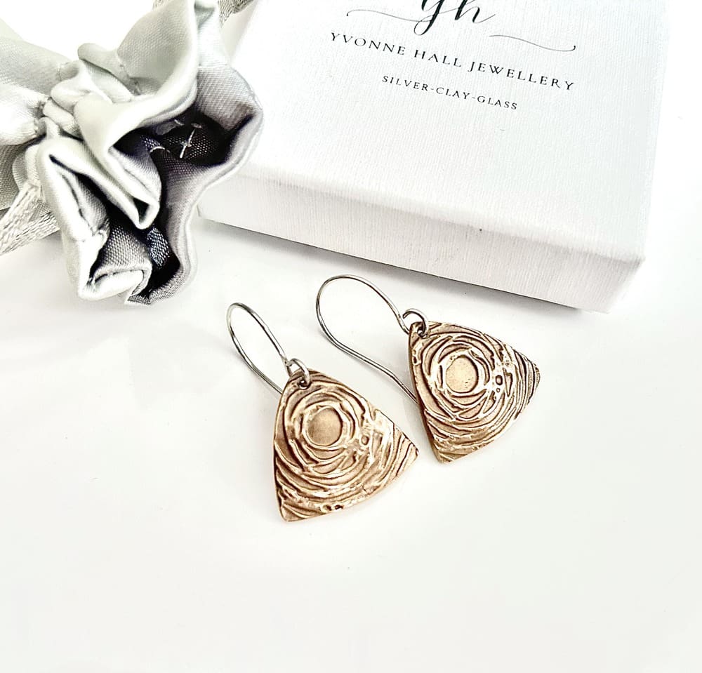 A pair of handcrafted bronze triangular drop earrings that have a swirl textured raised pattern. They have sterling silver ear wires and are displayed on a white gift box