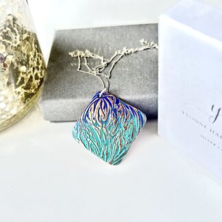 A handcrafted textured copper pendant which has a blue and green patina, it is in a diamond shape and has a sterling silver chain. The pendant is displayed on a white background with a grey gift box.