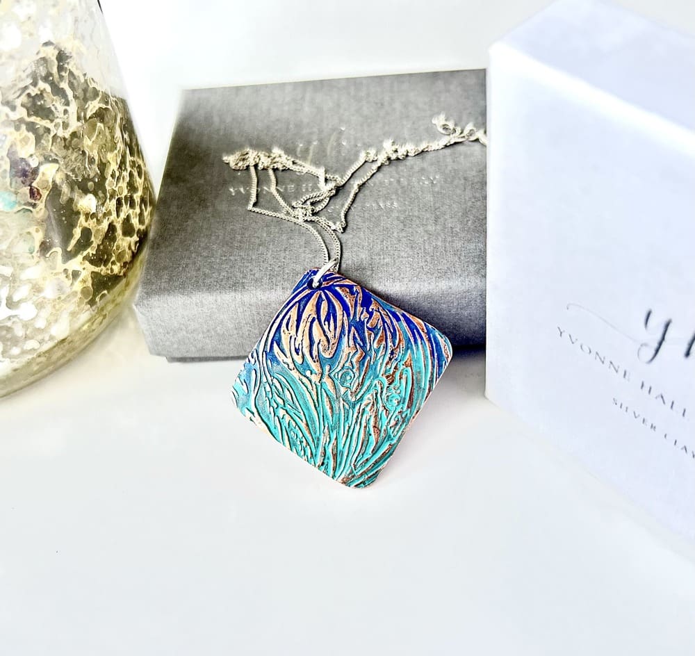 A handcrafted textured copper pendant which has a blue and green patina, it is in a diamond shape and has a sterling silver chain. The pendant is displayed on a white background with a grey gift box.