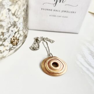 A round bronze pendant with a raised circular pattern and a hole in the centre. It has a sterling silver chain and is displayed on a white surface with acwhite gift box standing behind it.