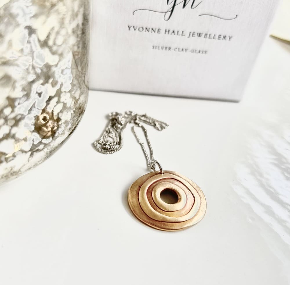 A round bronze pendant with a raised circular pattern and a hole in the centre. It has a sterling silver chain and is displayed on a white surface with acwhite gift box standing behind it.