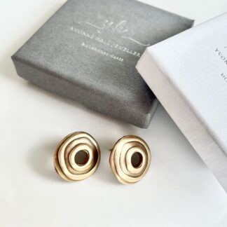 A pair of handcrafted bronze rounc stud earrings with a hole in the centre. They are displayed on a white surface with a grey and a white gift box above them.