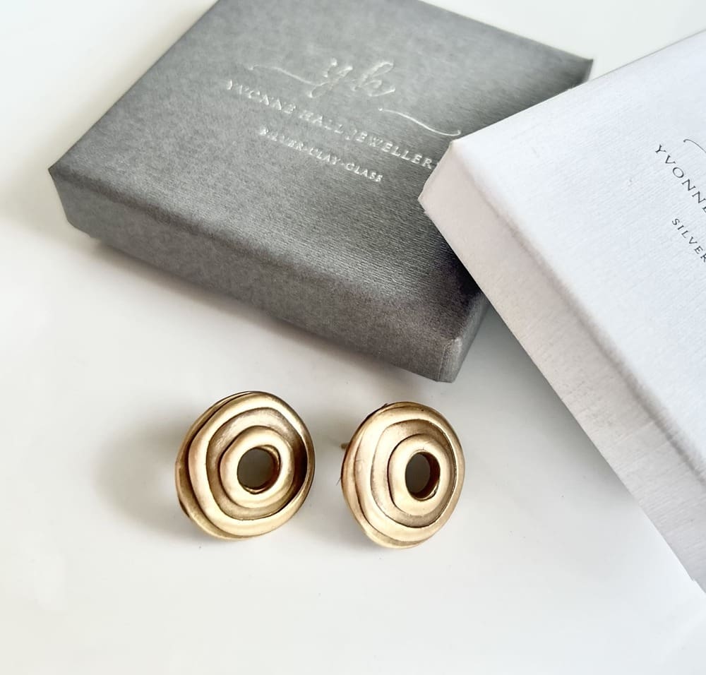A pair of handcrafted bronze rounc stud earrings with a hole in the centre. They are displayed on a white surface with a grey and a white gift box above them.