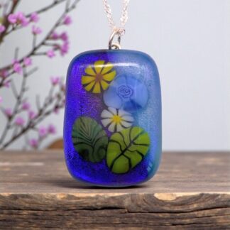 A vibrant blue glass pendant featuring colorful floral designs and green leaves, on a wooden surface with delicate pink blossoms in the background.