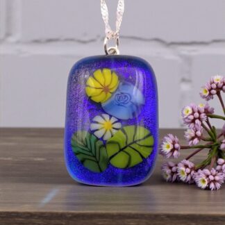 A vibrant glass pendant with floral designs is displayed on a wooden surface. The pendant features yellow, white, and blue flowers with green leaves, set against a deep blue background. A small cluster of purple flowers is positioned to the right, enhancing the delicate and artistic tone.