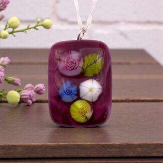 Cranberry coloured glass pendant featuring colorful floral designs, hanging on a silver cord, set against a wooden background with delicate flowers nearby.