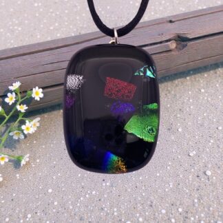 A dark dichroic glass pendant with colorful iridescent fragments, hanging on a cord against a textured background, with small white flowers nearby.