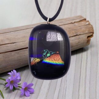 A dark dichroic glass pendant with iridescent patterns resembling mountains and a rainbow, resting on weathered wood and surrounded by purple flowers.